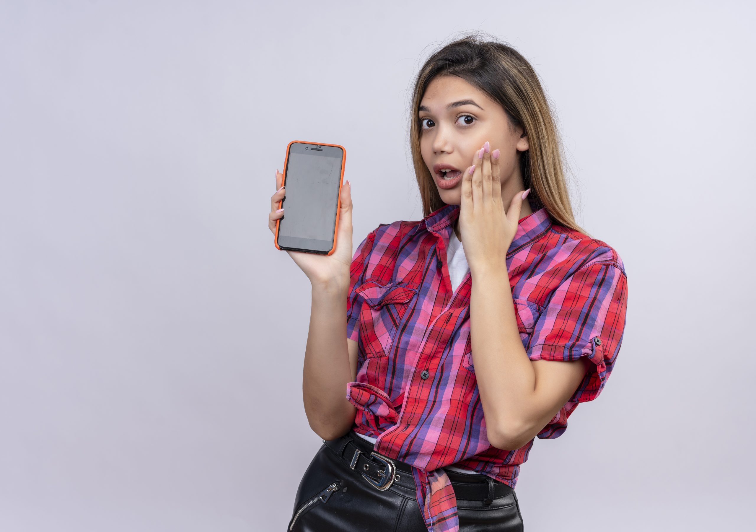 a surprised young woman in a checked shirt showing mobile phone while looking at the camera on a white background